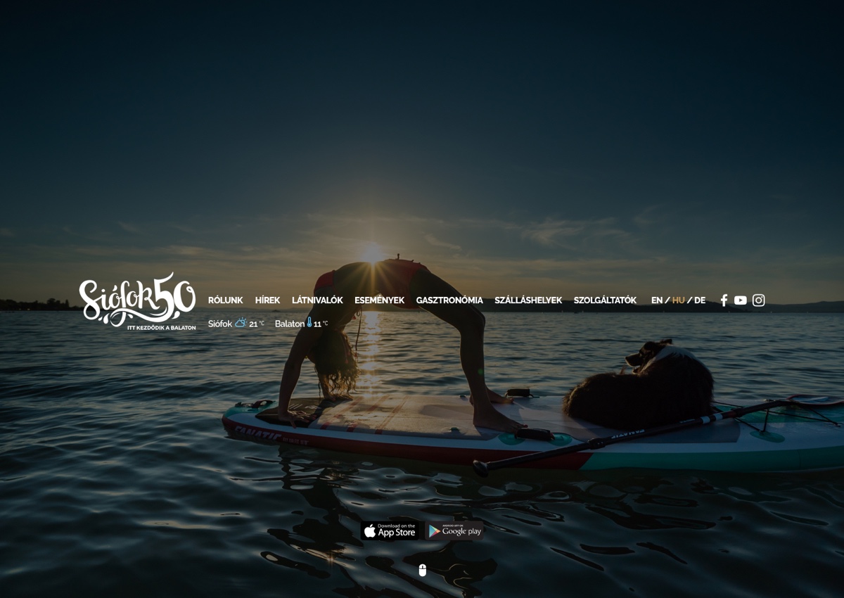 An earlier hellosiofok.hu homepage: a woman paddleboarding with her dog on Lake Balaton at sunset, with the site menu and live temperature readings for the town and the lake in the middle of the page, and App Store / Google Play download buttons near the bottom